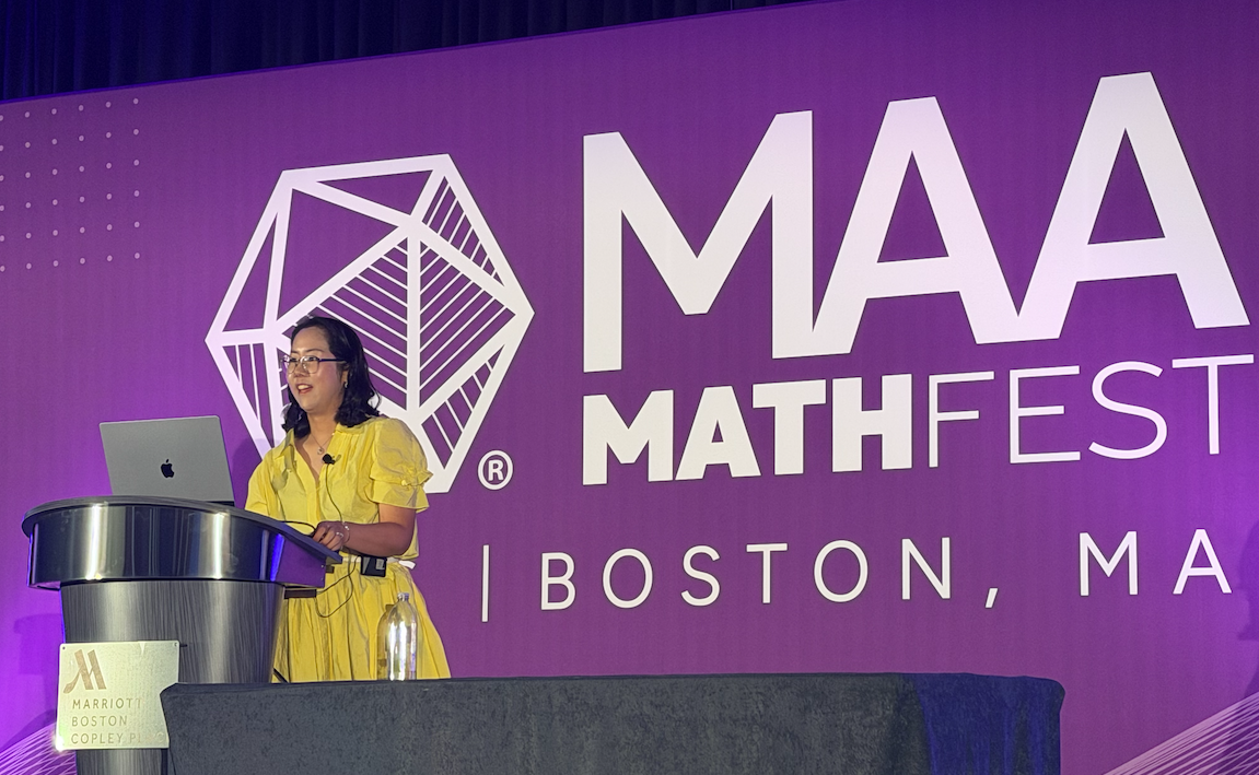 Hwayeon Ryu in a yellow dress standing at the podium presenting her keynote address at the MAA MathFest.
